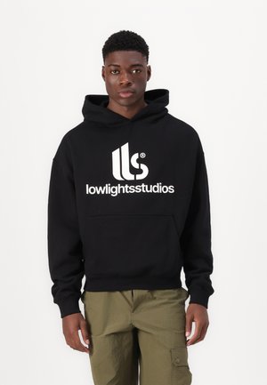 Čierna mikina s kapucňou a veľkým bielym logom "lowlightsstudios" na prednej strane, s prednou vreckom a šnúrkou na kapucni. Nosená s olivovozelenými cargo nohavicami.