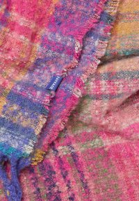 Multicolored plaid blanket in pink, purple, and yellow with a soft, textured surface and a fringed edge, featuring a blue fabric label.