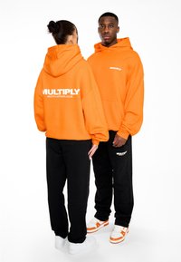 Sudaderas naranjas con texto blanco "MULTIPLY APPAREL CLUB", combinadas con pantalones negros. Las sudaderas tienen un corte relajado y bolsillos tipo canguro.