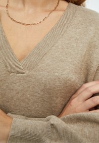 Bež pulover s V-izrezom napravljen od mekog materijala s rebrastom teksturom. Ima opušteni kroj i duge rukave, a dodatak mu je jednostavna lančić ogrlica.