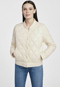 Pikowana kremowa kurtka z okrągłym kołnierzem, zatrzaskami i bocznymi kieszeniami. Miękka tekstura z prążkowanymi mankietami i dołem, w komplecie z niebieskimi dżinsami.