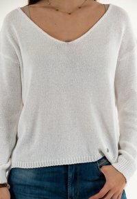 Suéter blanco de punto con escote en V, ajuste holgado y mangas largas. Presenta un dobladillo acanalado y un sutil logo de la marca en el lado inferior.