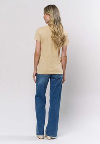 Béžové bavlněné tričko s krátkými rukávy, kombinované s světle modrými širokými džíny. Hladká textura, volný střih, nošeno s béžovými botami.