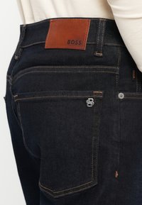 Tmavomodré džínsy z denimu s hnedou koženou nášivkou s vyrazeným nápisom "BOSS" na páse. Obsahujú kontrastné prešívanie a kovový logový akcent.
