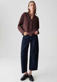 Woman standing with hands in pockets, wearing a loose brown long-sleeve blouse, dark wide-leg jeans, and black loafers on a plain background.