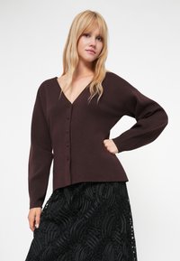 Cardigan lavorato a maglia marrone scuro con scollo a V, chiusura a bottoni e maniche lunghe, abbinato a una gonna nera strutturata con motivi ondulati.