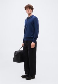 Νέος άνδρας με σγουρά μαλλιά, φοράει μπλε πόλο με μακριά μανίκια, μαύρο φαρδύ παντελόνι, μαύρα παπούτσια και κρατάει μια μαύρη τσάντα στο πλάι του.