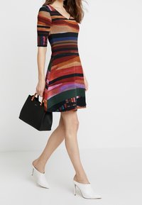 Abito a maniche corte con scollatura a V, caratterizzato da righe verticali arancioni, nere, verdi e viola. Indossato con una borsa nera e mule bianche con tacco.