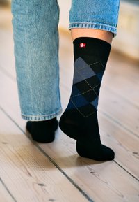 Calcetines negros con un patrón de argyle azul y acentos amarillos, rematados con un pequeño logotipo de bandera danesa en el puño.