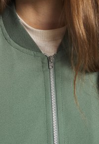 Zelená bomber bunda na zip s žebrovaným límcem a manžetami, s lesklým stříbrným zipem a hladkou texturou látky. Světle béžová vnitřní vrstva je viditelná.