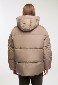 Giubbotto insolato in marrone chiaro con cappuccio. Presenta un design trapuntato e imbottito, con una consistenza spessa e morbida che crea una silhouette voluminosa.