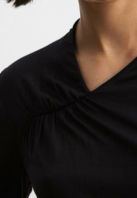 Top nero sagomato con scollatura a V e dettagli arricciati lungo il lato, realizzato in tessuto leggero e liscio.