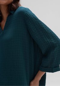 Błękitna bluzka o fakturze z wzorem w kratkę, luźny fason, kołnierzyk koszulowy i podwinięte rękawy o długości trzech czwartych. Miękka tkanina z swobodnym krojem.