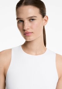 Vit ärmlös topp med hög hals och slät textur, med en subtil sömdetalj i mitten. Modellen har rakt hår.