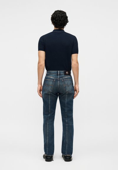 Bărbat cu păr creț, purtând o cămașă bleumarin cu mâneci scurte, blugi albaștri decolorați și adidași negri, stând cu spatele către cameră pe un fundal simplu.