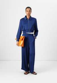 Veste courte bleu marine avec fermeture éclair, taille élastique et pantalon large assorti. Tient un sac à main orange vif. Sandales plates.