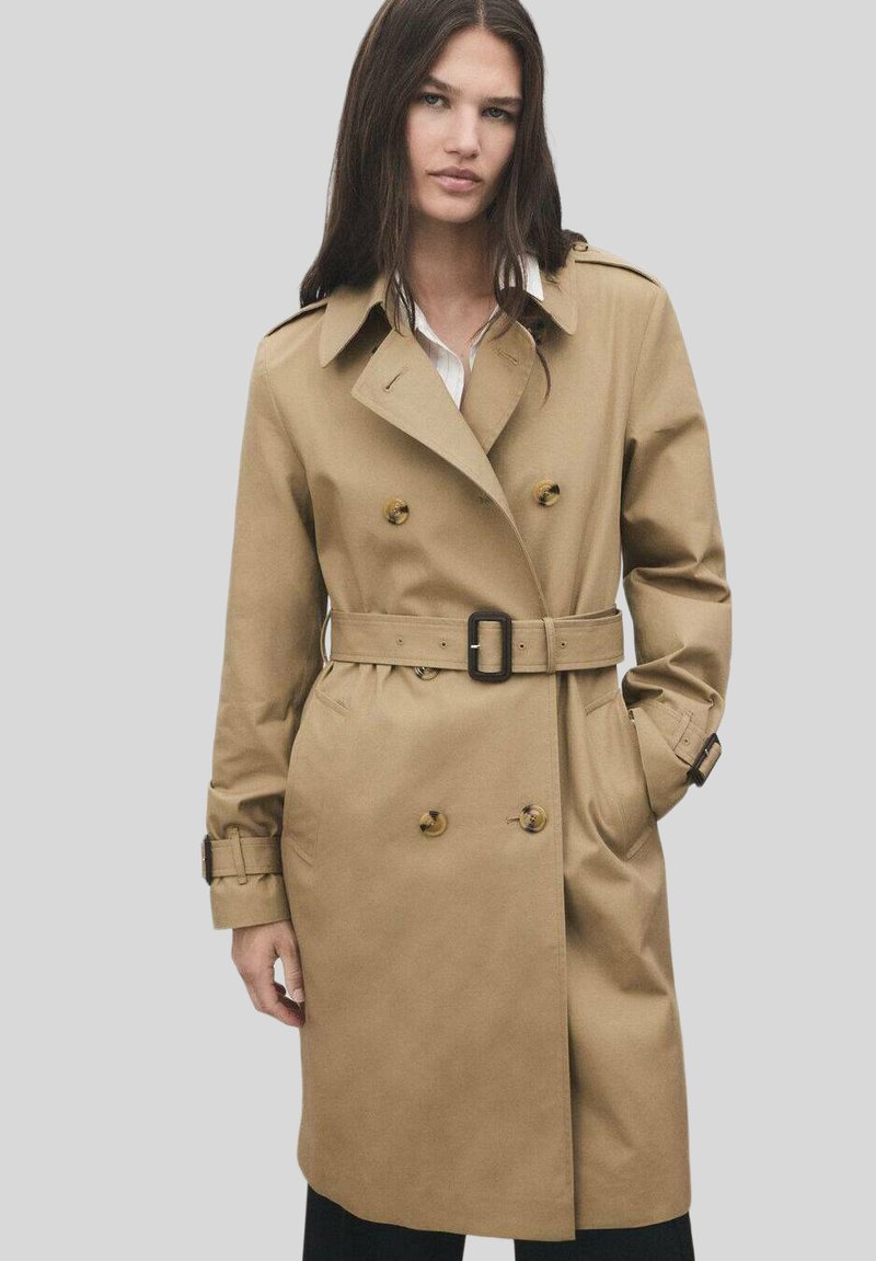 Trench coat beige con design doppiopetto, ampio colletto, vita con cintura, dettagli con bottoni e cinturini ai polsini; il tessuto sembra liscio e leggero.
