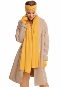 Écharpe jaune, bandeau et gants en tissu doux ; manteau beige au design ajusté avec de larges revers, présentant un détail de bouton.