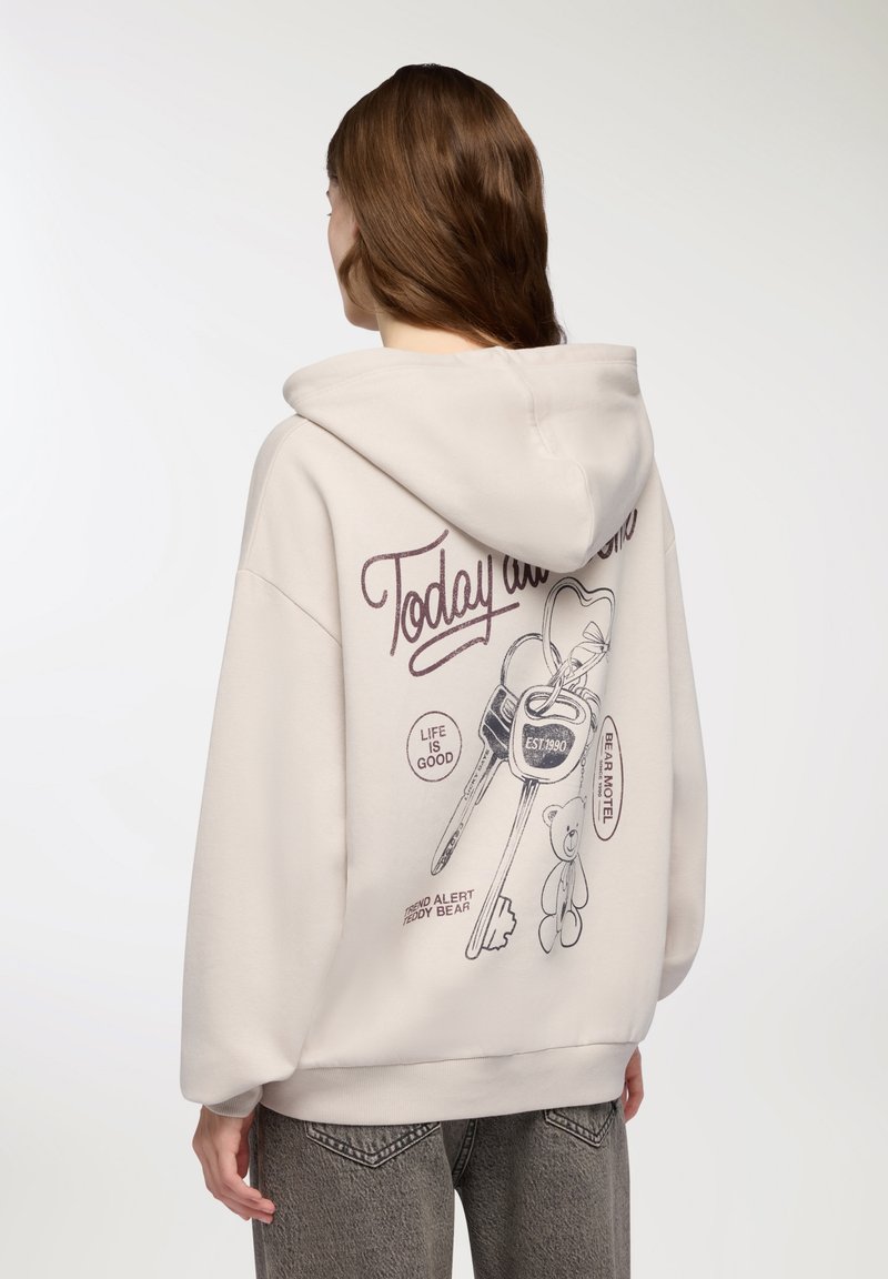 Beżowy hoodie z kieszenią kangurką. Na plecach graficzny wzór przedstawiający klucze, misia pluszowego i tekst. Wykonany z miękkiego materiału; luźny krój.