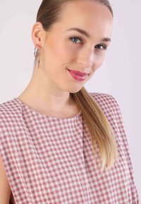 Die blusige Bluse hat ein pink-weißes Karomuster, besteht aus leichtem Stoff und hat eine lockere Passform. Das Model trägt Ohrringe mit Creolen.