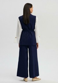Mörkblå linné väst med b�älte, kombinerad med vida mörkblå byxor. Vit långärmad skjorta med manschettdetaljer. Minimalistisk design.