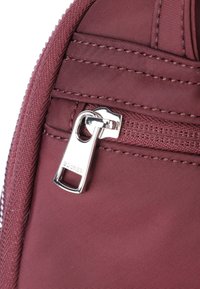 Burgundy tygväska med ett silverfärgat dragkedjehandtag. Dragkedjeleden är synlig och har texturerad sömnad samt en logotyp ingraverad på metallföremålet.