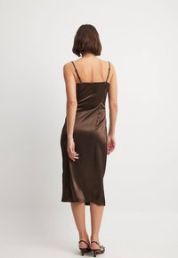 Brązowa satynowa sukienka na ramiączkach, dopasowana sylwetka, długość do połowy łydki, gładka tekstura i subtelny połysk. Czarne sandały na obcasie.