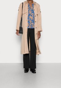 Trench-coat beige, blouse à motifs bleus et orange, pantalon large noir, et chaussures noires. Petit sac bandoulière noir.