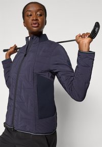 Veste puffer marine avec un col haut, dotée d'un panneau latéral en maille texturée et d'une fermeture éclair. Des accents noirs soulignent les poignets et l'ourlet.
