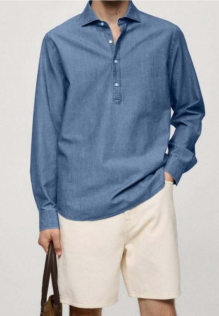 Homme portant une chemise bleue à manches longues boutonnée et un short beige, tenant un sac à main marron dans une main, debout devant un fond uni.
