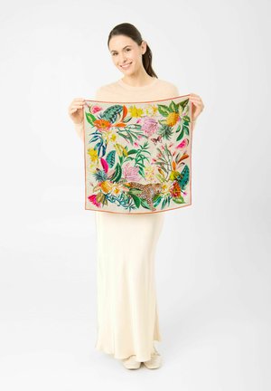 Femme en robe crème tenant un foulard carré avec un imprimé floral tropical et animal, incluant des ananas, des feuilles et un léopard.