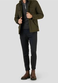 Giacca verde oliva con zip e due tasche frontali, indossata sopra un maglione grigio. Pantaloni scuri e strutturati e stivaletti marroni alla caviglia.