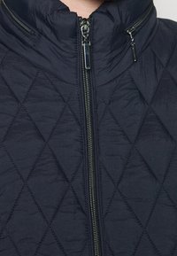 Chaqueta acolchada azul marino con cuello alto, que cuenta con una cremallera central y dos bolsillos laterales con cremalleras. La textura parece suave y ligera.