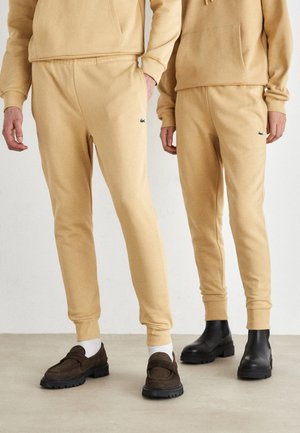 Pantalones de chándal beiges con cintura y puños elásticos, con un pequeño logo verde en el lado izquierdo. Combinados con zapatos oscuros contrastantes.