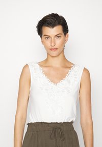 Top branco sem mangas com decote texturizado em bordado floral, apresentando um formato em V profundo. Combinado com calças verde-azeitona com cordão na cintura.