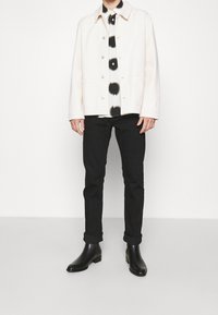 Weiße Jacke mit Knöpfen über einem schwarz-weißen gemusterten Hemd, kombiniert mit hochgekrempelten schwarzen Hosen und schwarzen Stiefeletten.