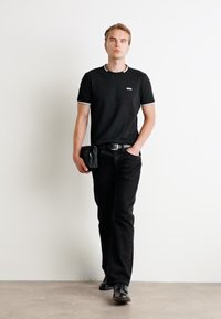 Sort T-shirt med hvide kraveakcenter, båret med sorte jeans. Et sort bælte og en lille sort taske er synlige. Modellen står foran en hvid væg.