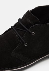 Chaussure à lacets en suède noir avec œillets métalliques et coutures visibles sur la semelle, montrant une partie de la tige et de la languette.
