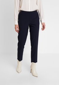 Marina skräddarsydda byxor med en slät yta, har sidofickor och silverdetaljer vid midjan, som är ihop med krämfärgade högklackade stövlar.