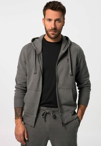 Sudadera gris con cremallera y bolsillo canguro, puños acanalados y capucha con cordón, llevada sobre una camiseta negra y joggers grises a juego.