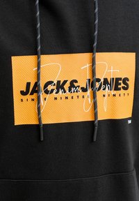Sweat à capuche noir avec un logo rectangulaire orange portant l'inscription "JACK & JONES Denim People Since Nineteen Ninety" et des cordons noirs à pois blancs.