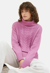 Pulover roz cu tricotaj de cabluri, cu guler înalt, având mansete cu margine ondulată și o croială confortabilă. Modelul îl poartă cu pantaloni albi.