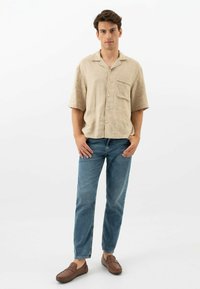 Camicia di lino beige chiaro a maniche corte con bottoni e taschino, abbinata a jeans blu e scarpe slip-on marroni, in piedi su uno sfondo bianco.