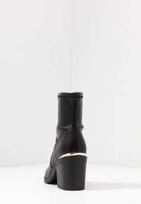 Bottine noire avec une texture en cuir lisse, de forme effilée et un talon épais. Présente un accent doré à la base du talon.