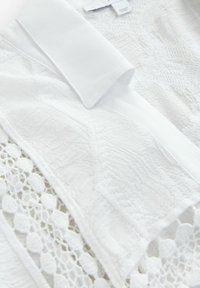 Vêtement blanc avec un tissu texturé au look froissé, un col uni et une garniture en dentelle le long des bords.