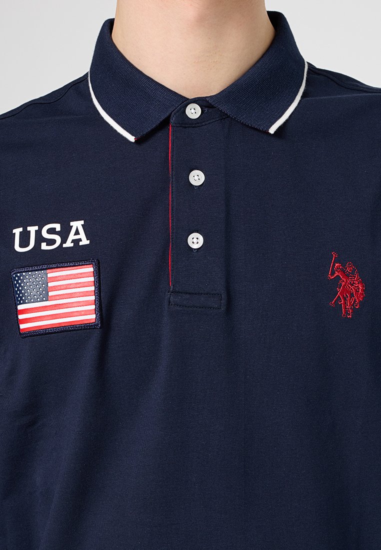 Polo de color azul marino con ribete blanco, que presenta un parche de EE. UU. y un logotipo bordado rojo en el pecho. Diseño de tapeta con cuatro botones.