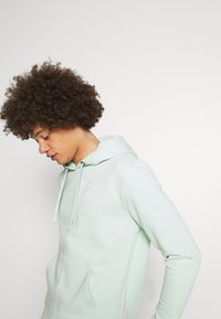 Hoodie verde claro feito de tecido suave, com um bolso frontal, capuz com cordão e um pequeno logo branco no peito.