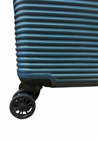 Valigia rigida di colore blu texturizzato, con un design a coste. Presenta quattro ruote nere e una struttura angolare robusta e arrotondata.