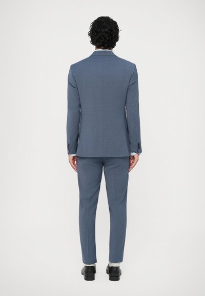 Sinine ülikond, mis on valmistatud täpselt sobivaks, ühe ventiiliga jakk ja kitsad püksid. Sile kangas ja mustad elegantejalatsid. Minimalistlik disain, ilma mustriteta.