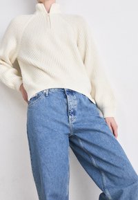 Krämfärgad ribbad stickad tröja med hög krage och halvdragkedja. Kombinationen med ljusblå, baggy jeans med kontrasterande sömmar och knäppning.